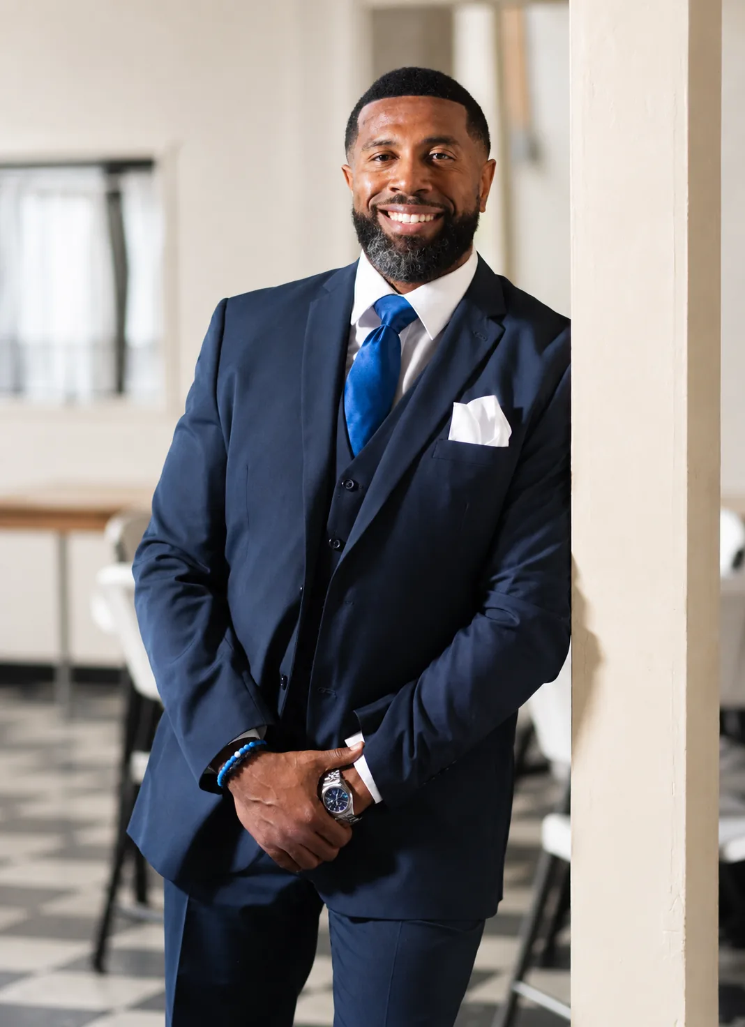 Cory J. Chavis, a sitting principal and school-culture speaker, smiling in a navy three-piece suit with a blue tie and white pocket square, leaning against a column.